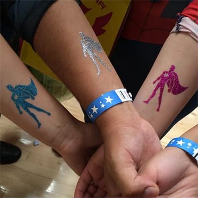 Three arms with glitter tattoos of superheroes and a wristband with stars.