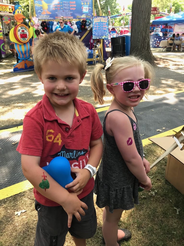 Two children at a fair, one holding a blue balloon and both sporting temporary tattoos.