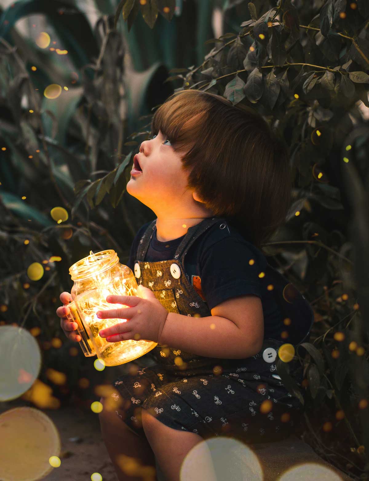 A child holding a jar of lights, gazing upward amidst a backdrop of leaves.