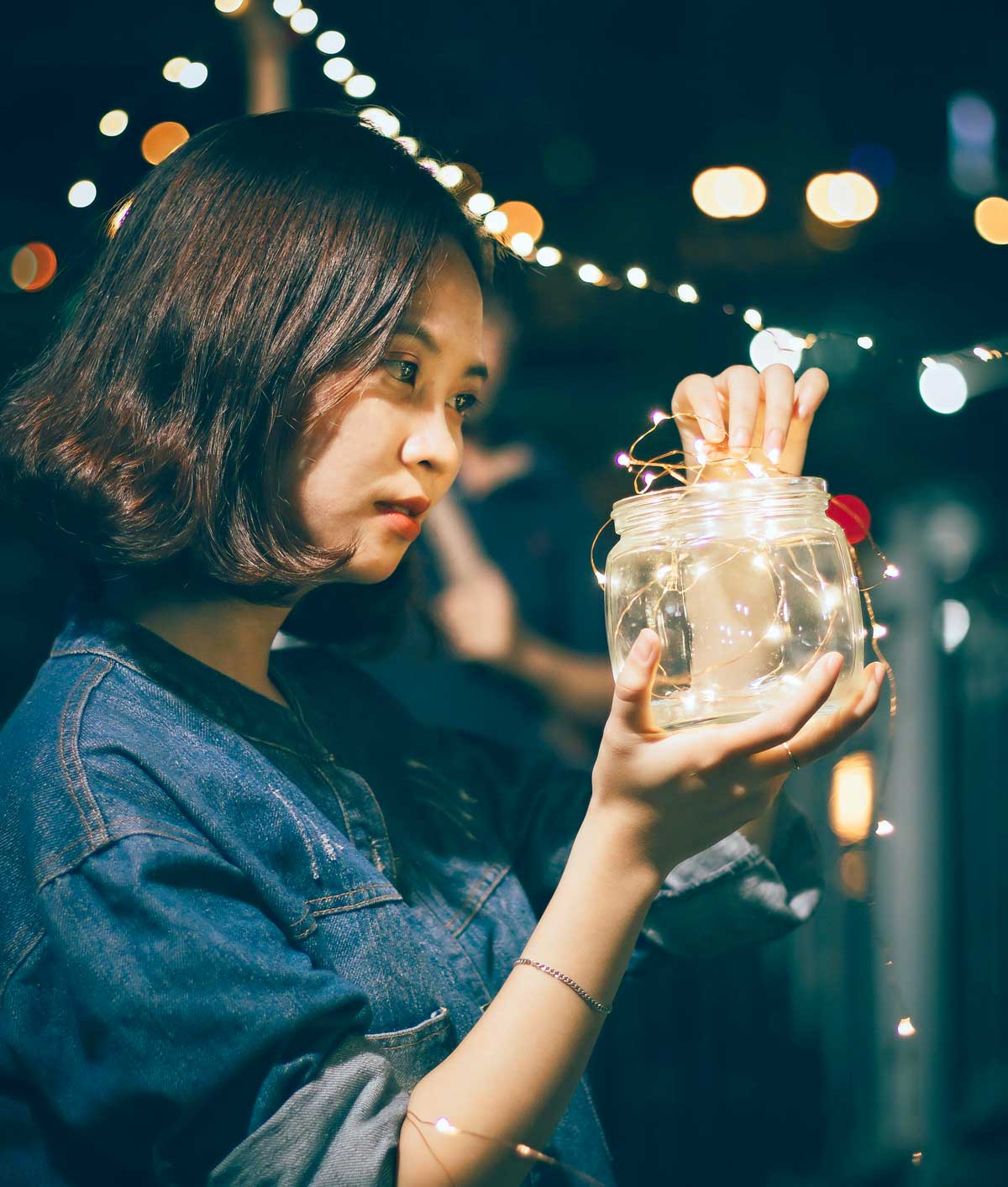 A woman holding a jar with lights inside, surrounded by a bokeh effect from additional lights in the background.
