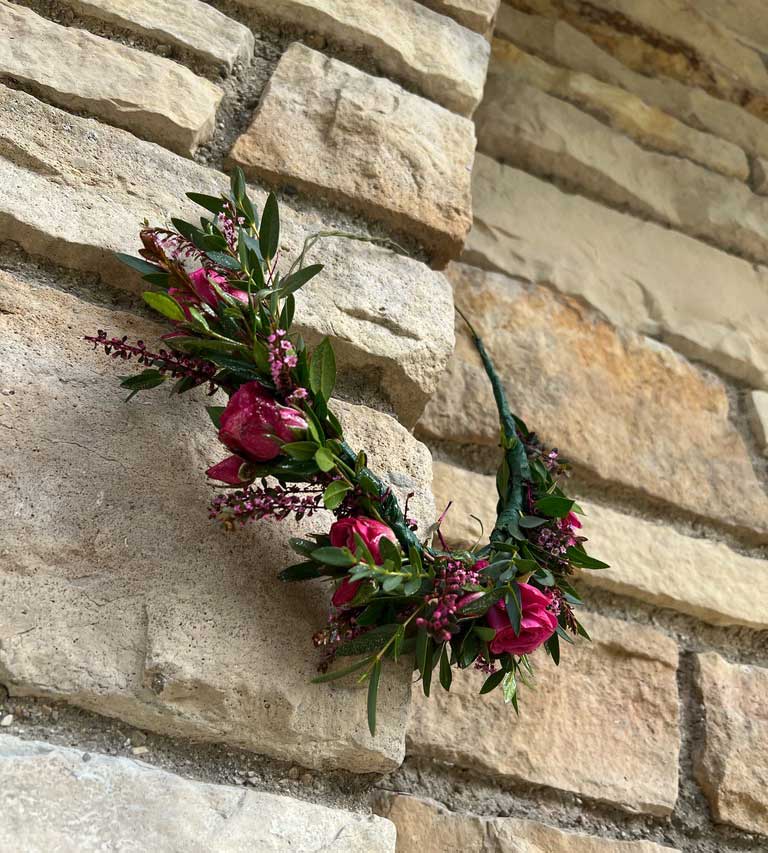 A floral wreath with pink flowers and green leaves hanging on a textured stone wall.