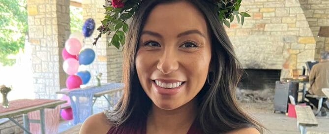 A smiling woman wearing a floral headpiece and a burgundy bridesmaid dress at an outdoor wedding with balloons in the background.