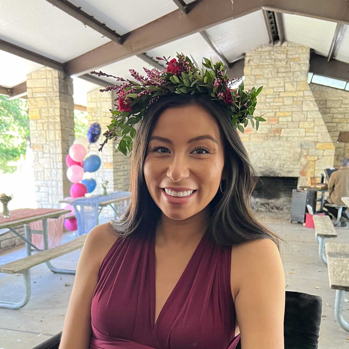 A smiling woman wearing a floral headpiece and a burgundy dress at an outdoor event with balloons in the background.