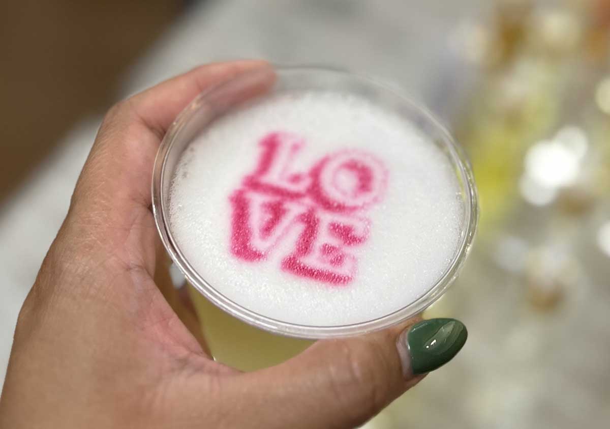 A hand holding a cup of latte with the word "love" stenciled in pink on the foam.