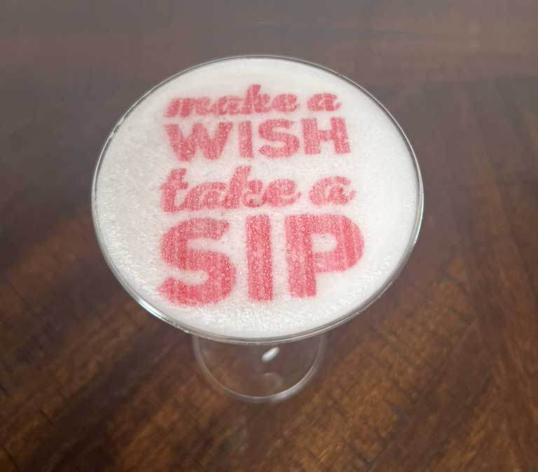 A frothy drink with the words "make a wish take a sip" sprinkled on top in red.