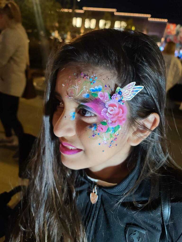 Woman with festive face paint including a pink rose, blue dots, and glitter at a night event.