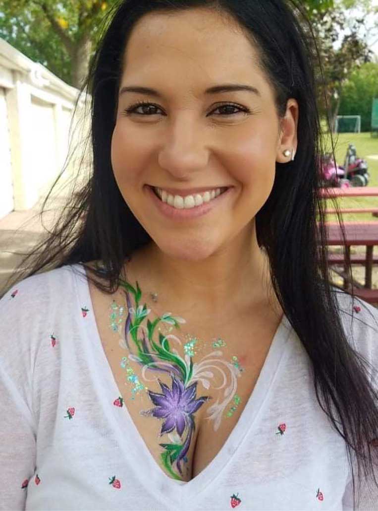 A woman with a painted flower on her chest smiling outdoors.