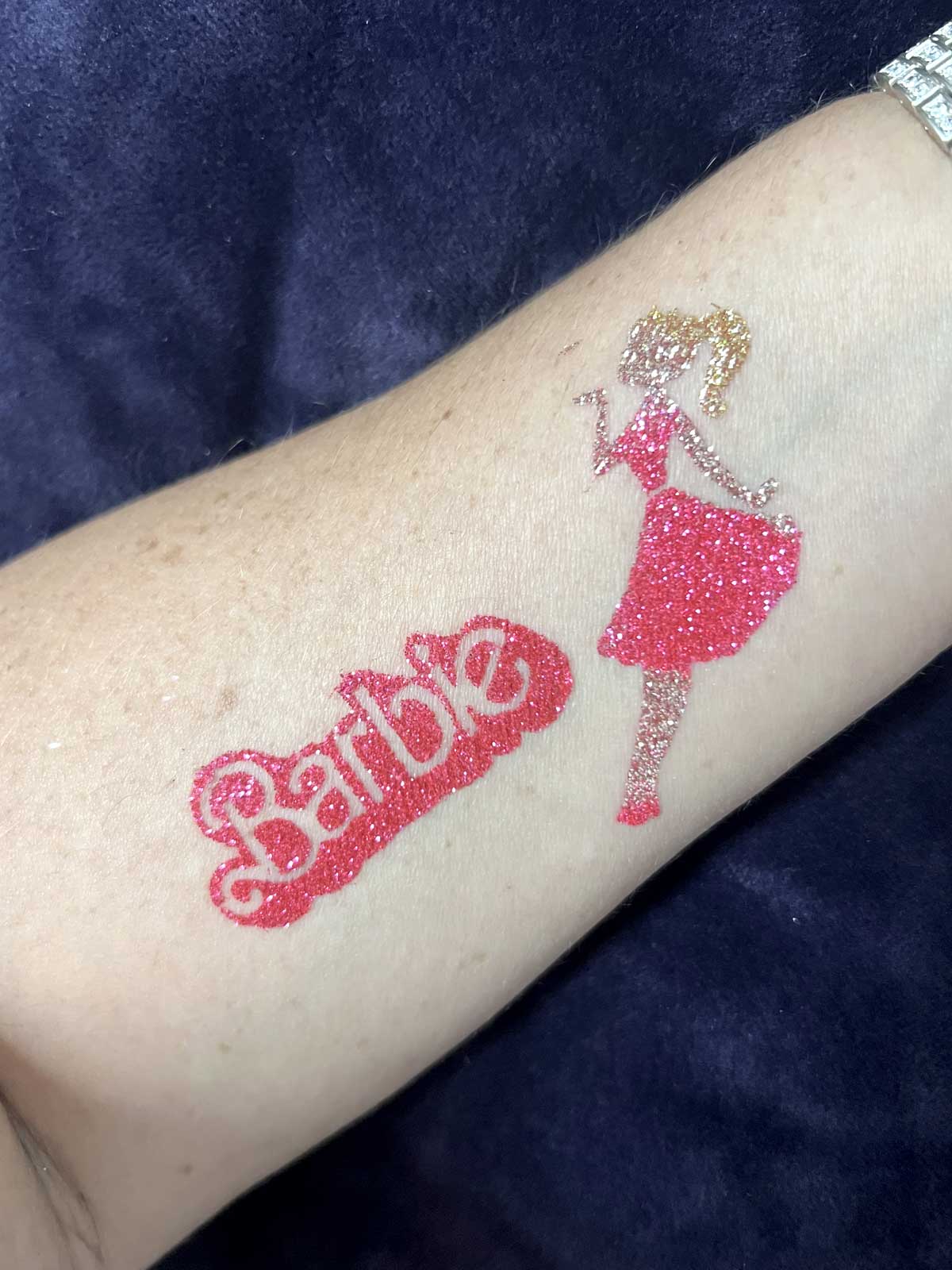 A sparkly pink temporary tattoo of the barbie logo and a silhouetted figure on a person's arm.