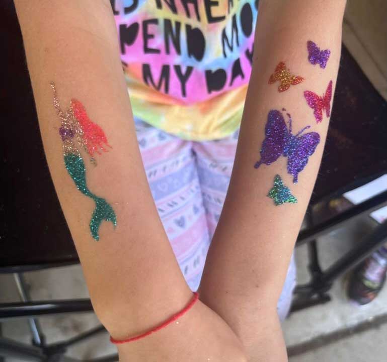 Child's arms with colorful glitter tattoos of a mermaid tail and butterflies.