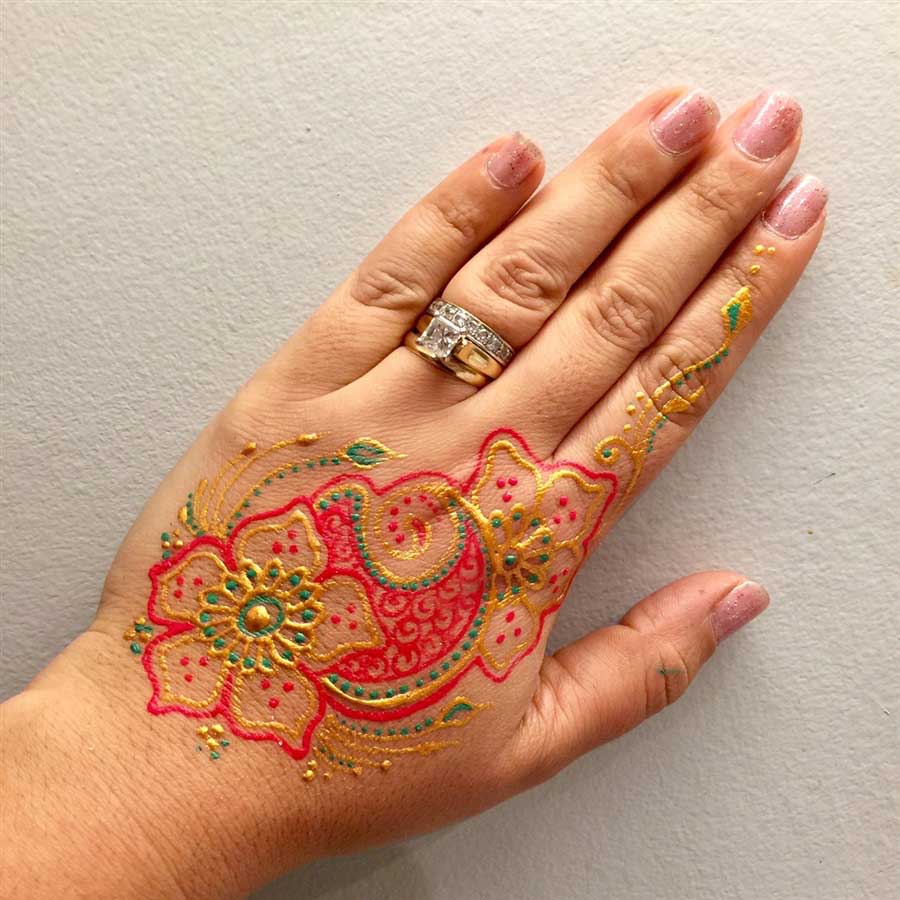 A hand displaying pink nail polish and a glittery pink and gold henna design, accessorized with a diamond ring.