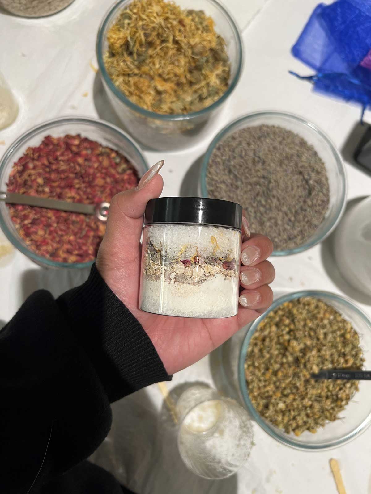 A person's hand holding a jar of layered spices with various bowls of herbs and spices in the background.