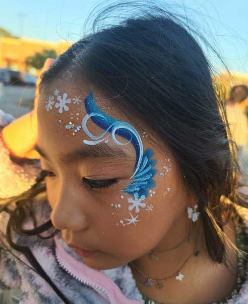 A child with a blue and white winter-themed face painting featuring snowflakes.