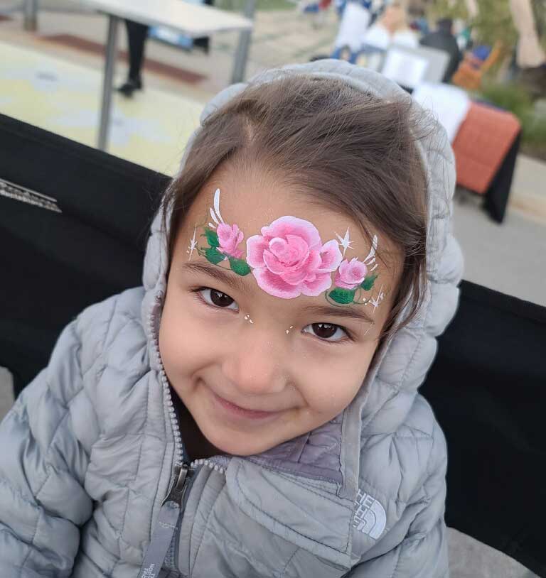 A young girl with face paint of pink flowers and glitter on her forehead smiling at the camera.