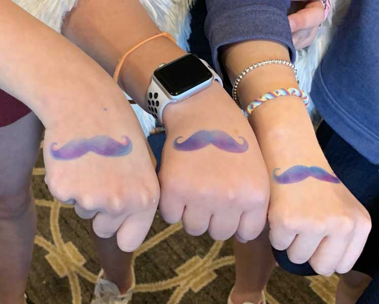 Four individuals displaying their wrists with matching mustache tattoos; one wrist wears a smartwatch.