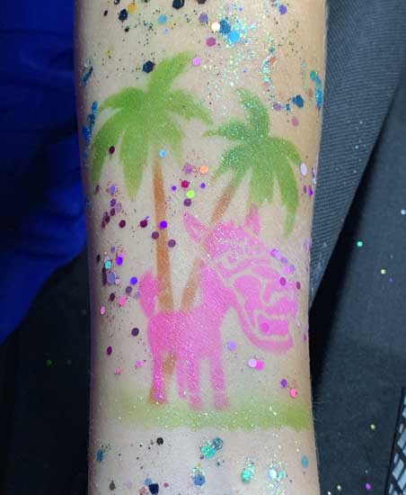 A glittery temporary tattoo of a pink unicorn under palm trees on someone's arm.