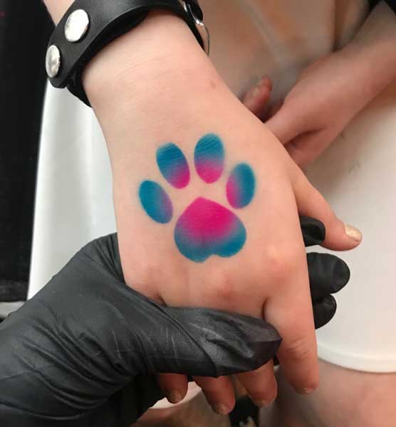 A person's hand displaying a freshly inked multicolored paw print tattoo, with a tattoo artist's gloved hand holding it.