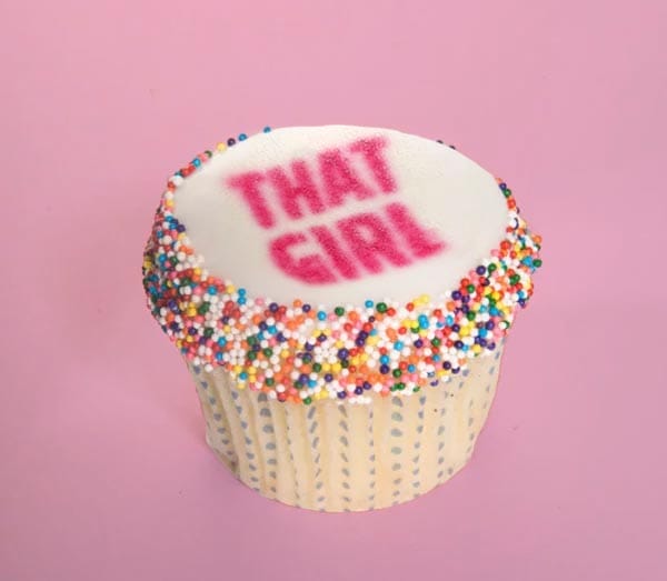 A cupcake with colorful sprinkles and "that girl" written on top, displayed against a pink background.