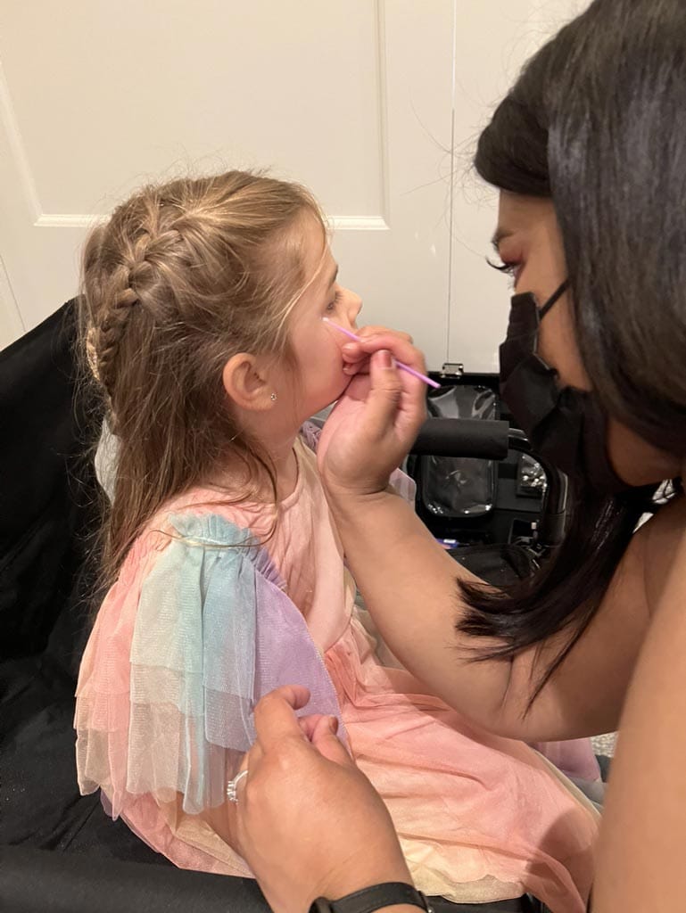 A child is seated and having makeup applied by an adult.