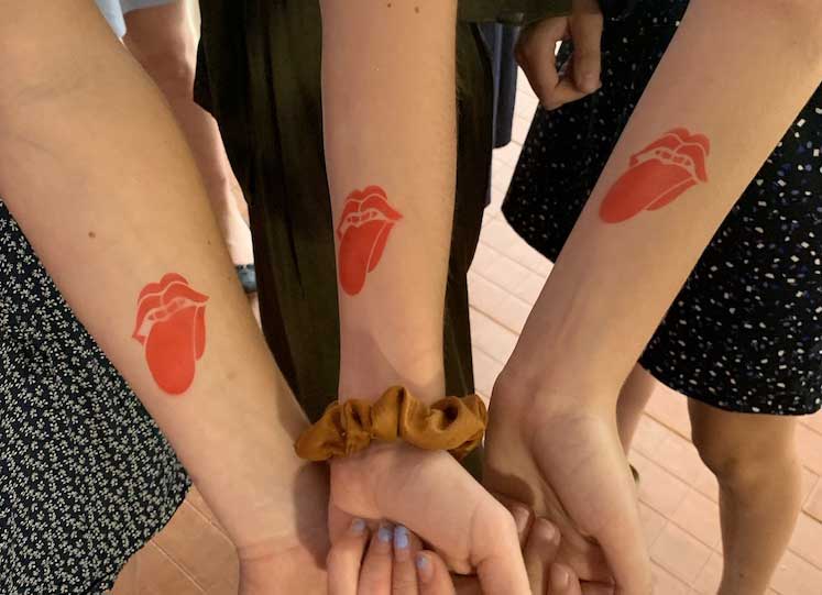 Four individuals showing their forearms with matching red heart tattoos, holding hands in a circle.