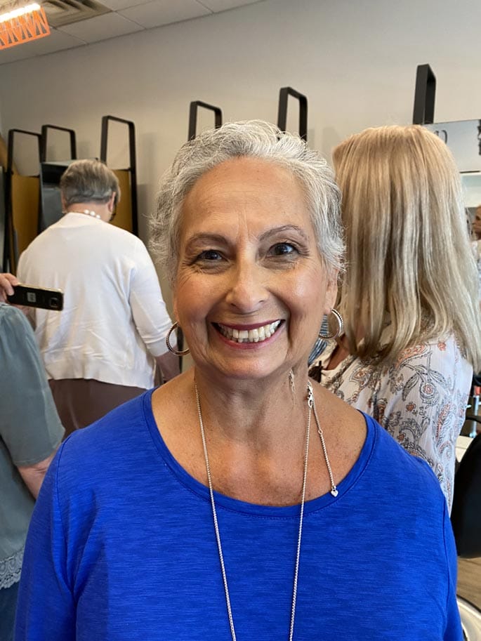A smiling woman with grey hair wearing a blue top and a silver necklace in a room with other people in the background.