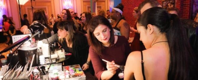 A face painting artist is preparing paint at an indoor wedding while bridal parties look on.