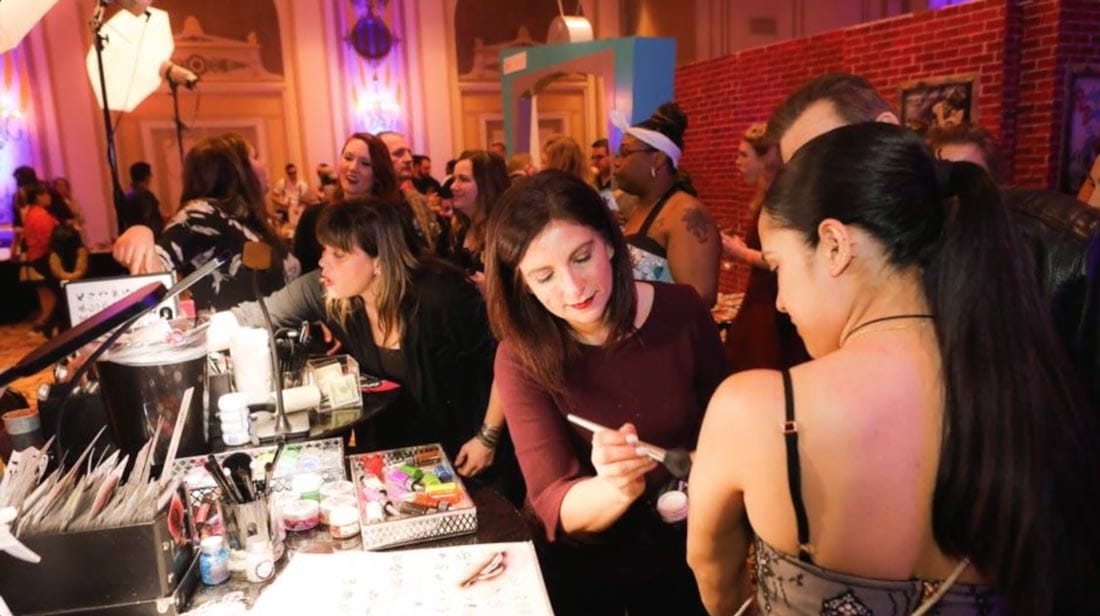 A face painting artist is preparing paint at an indoor wedding while bridal parties look on.
