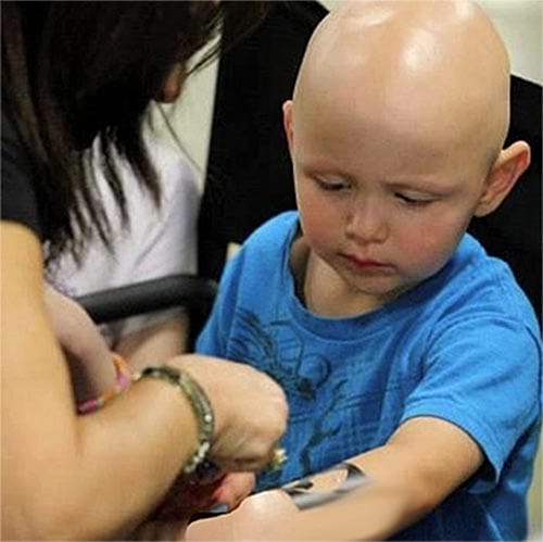 A young child with a bald head looking intently at a watch being put on his wrist by an adult.