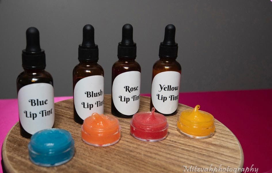 Assorted lip tint products with dropper bottles displayed alongside open containers of colored lip balm on a wooden holder.