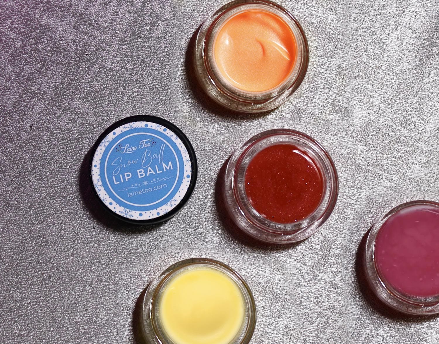 Various shades of lip balm in open jars arranged on a glittering surface.