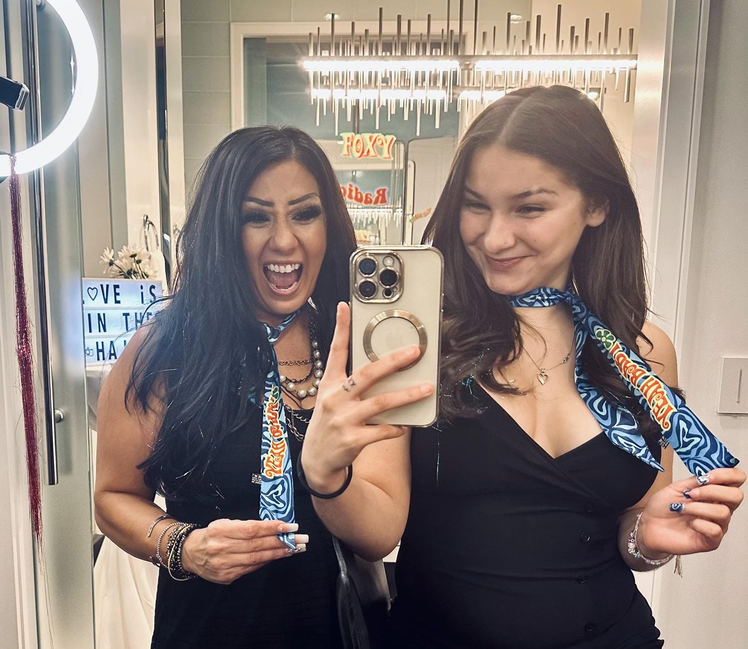Two women posing for a selfie in a mirror, one making a surprised expression and the other smiling, both wearing decorative scarves.