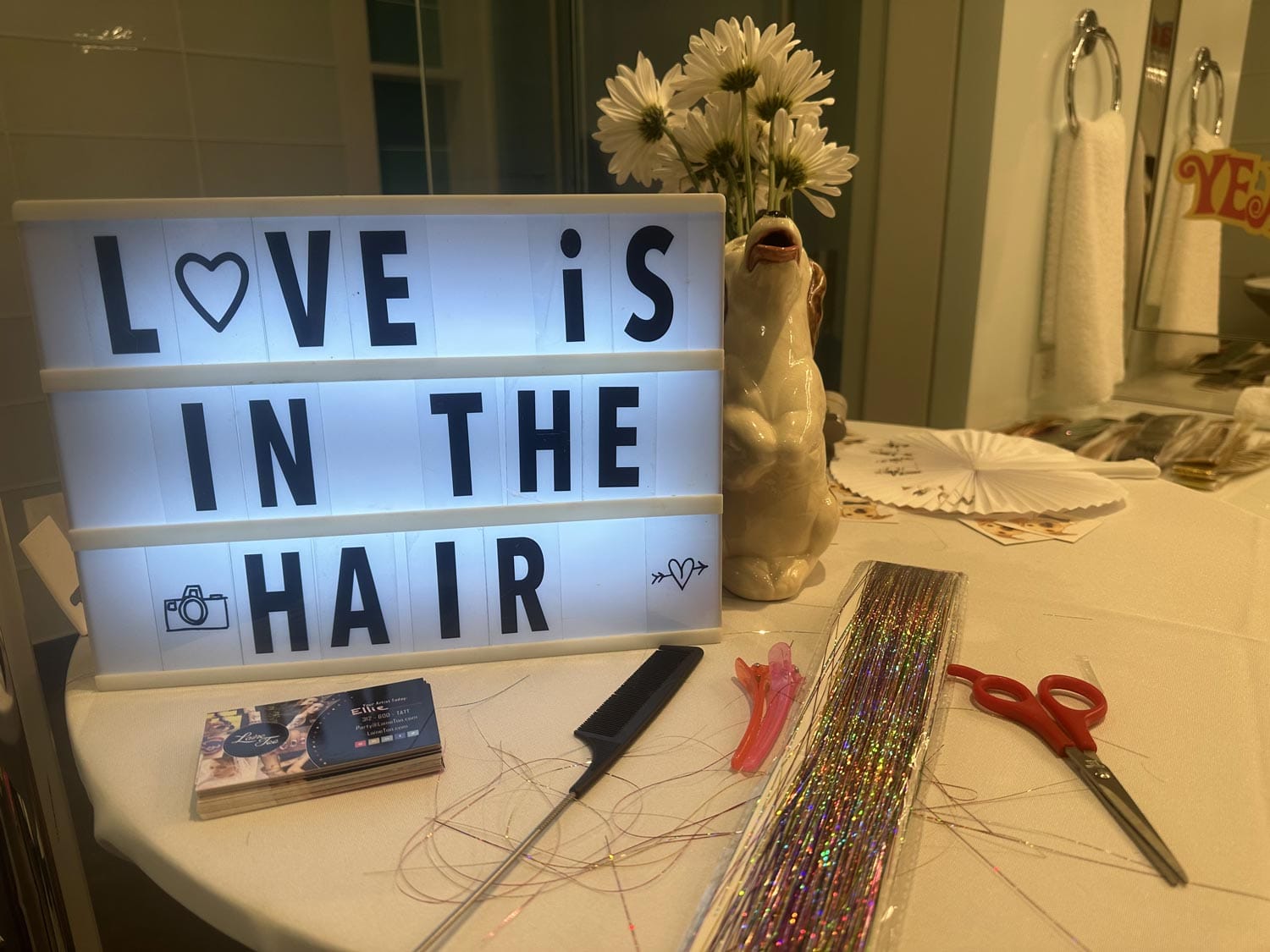Lightbox with the phrase "love is in the hair" on a table with a vase of white flowers, hair accessories, and scissors.