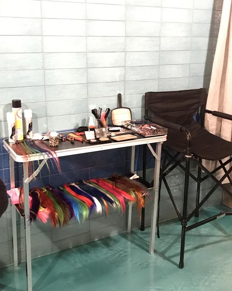 A makeup table with various cosmetics and brushes, next to a folding chair and a colorful costume hanging underneath, set against a tiled wall.